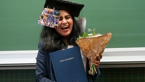 Eine Frau mit doktorhut lacht und hält ein Buch mit der Aufschrigt Doktorarbeit sowie einen Blumenstrauß