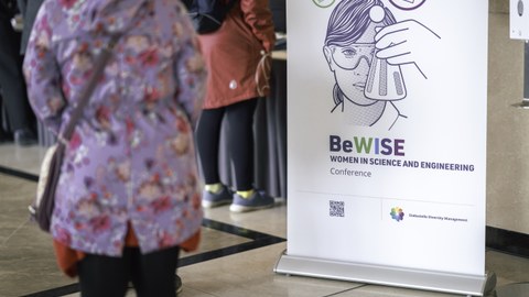 Person in floraler Jacke steht vor einem Poster auf dem eine Frau abgebildet ist, die einen Erlenmeyerkolben hält. Auf dem Poster steht "BeWISE Women in science and engeneering conference".