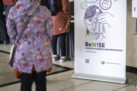 Person in floraler Jacke steht vor einem Poster auf dem eine Frau abgebildet ist, die einen Erlenmeyerkolben hält. Auf dem Poster steht "BeWISE Women in science and engeneering conference".