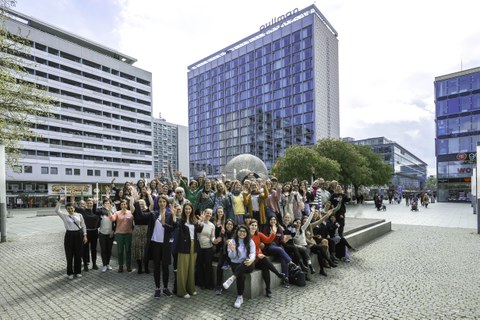 Eine Gruppe von Frauen steht und sitzt vor einem Pullman Hotel und hebt die Arme.