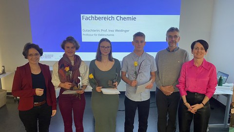 Zwei Frauen und ein Mann stehen neben zweiFrauen und einem Mann, die jeweils eine gelbe Rose und ein Papier in der Hand halten. Im Hintergrunf ist eine Präsentation zu sehen auf welcher Fachbereich Chemie steht.