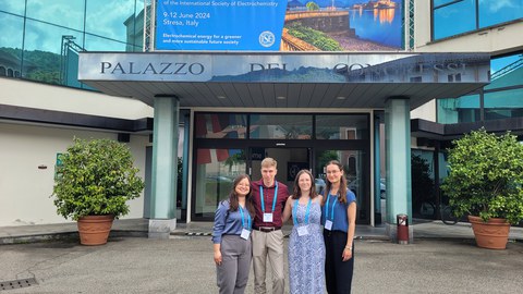 Vier Personen stehen vor einem Gebäude an dem ein Schild hängt auf dem   37th Topical Meeting  of the International Society of Electrochemistry steht.