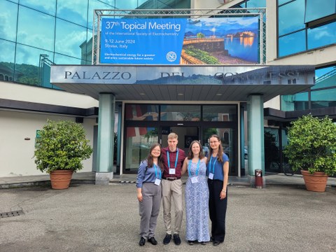 Vier Personen stehen vor einem Gebäude an dem ein Schild hängt auf dem   37th Topical Meeting  of the International Society of Electrochemistry steht.