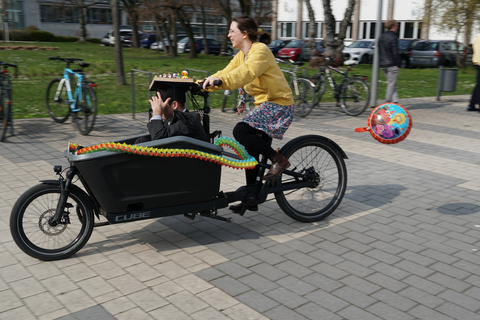 Eine Frau fährt ein Lastenrad in dem ein Mann mit Doktorhut vornne drin sitzt. 