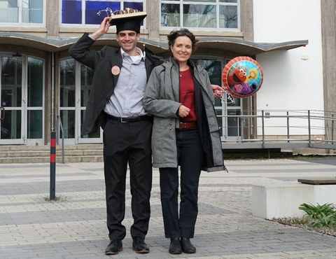Ein Mann mit Doktorhut steht neben einer Frau vor einem Gebäude, neben ihenen ein Ballon der Sesamstraße.