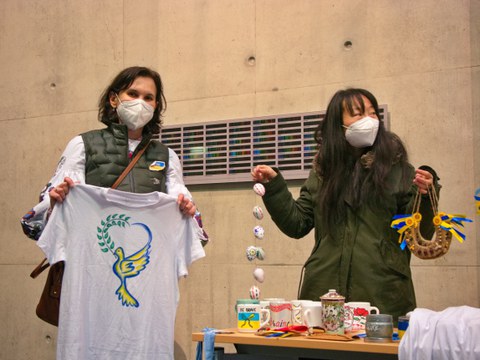 Zwei Frauen an einem Tisch mit verschiedenen Gegenständen zum Verkauf. Eine Frau hält ein weißes T-Shirt mit zwei Händen hoch. Auf dem T-Shirt ist eine stilisierte, gelb-blaue Friedenstaube abgebildet. Die zweite Frau hält eine Kette mit bunt bemalten Eiern sowie zwei verzierte Hufeisen hoch.