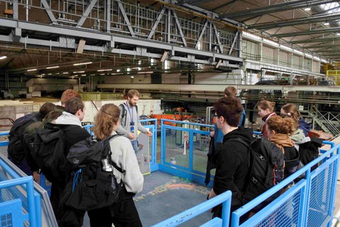 Eine Gruppe von Studierenden mit einem Führer steht auf einer Aussichtsplattform in einer Halle mit Experimenten.