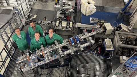 Blick von oben in eine Halle mit experimentellen Aufbauten, davor 4 Personen in grünen Arbeitskitteln