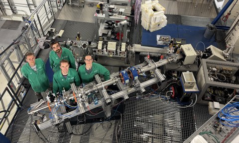 Blick von oben in eine Halle mit experimentellen Aufbauten, davor 4 Personen in grünen Arbeitskitteln