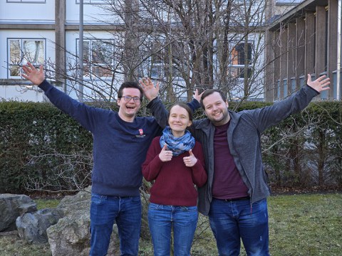 A group of three people; two men raise their arms in the air, the woman in the middle points both thumbs up