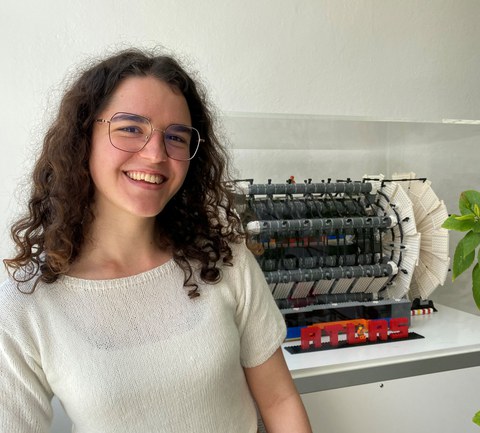 Eine junge Frau mit Brille, schwarzen Locken und weißem Pulli steht vor einem Lego-Modell des ATLAS-Detektors