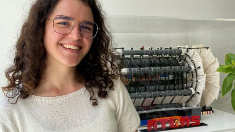 Eine junge Frau mit Brille, schwarzen Locken und weißem Pulli steht vor einem Lego-Modell des ATLAS-Detektors