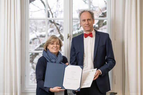 Zwei Personen stehen vor einem Fenster und halten geöffnete Mappen mit Urkunden; winterliche Landschaft im Hintergrund.