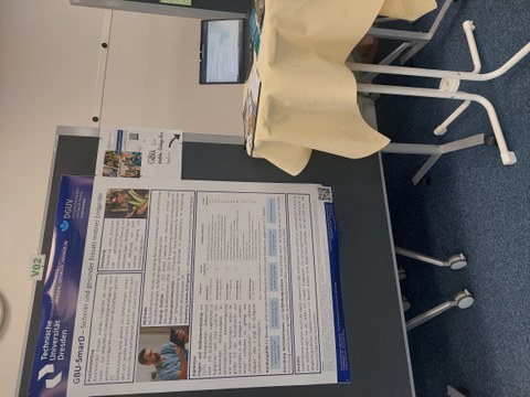 Ein wissenschaftliches Poster auf einer Pinnwand. Daneben ein Beistelltisch mit Laptop. 