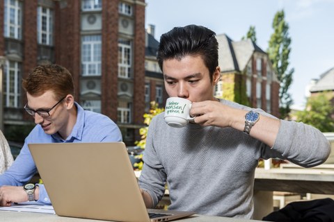 Zwei Männer lernen im Freien. Einer davon trinkt aus einer Tasse mit "Lieber mensen gehen!" drauf.