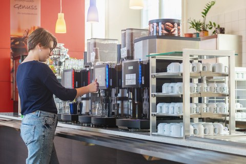 Das Foto zeigt eine Studentin in der Mensa. Sie steht gerade an einer der drei Kaffeeautomaten. Im Vordergrund sieht man viele Kaffeetassen.
