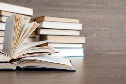 Auf dem Foto liegen zwei aufgeschlagene Bücher auf einem Tisch. Im Hintergrund ist ein Bücherstapel zu sehen