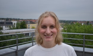 Portraitaufnahme von Beatrix von Woedtke. Eine Frau mit heller Haut und mittellangen blonden Haaren steht auf einer Dachterrasse und lächelt in die Kamera.