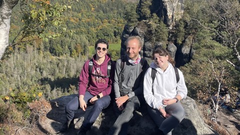 Kristina Rainich, Gael Cordero Otero und Jonathan Wehnert sitzen auf einem Berggipfel in der Sächsischen Schweiz.