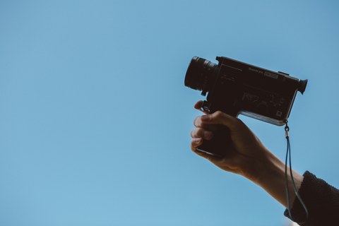 Eine Hand hält eine Videokamera, im Hintergrund sieht man blauen Himmel.