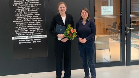 Dr. Sofia Meyer und Prof. Katharina von Kriegstein stehen nebeneinander vor einem Vorlesungsraum, Sofia hat einen Blumenstrauß in der den Händen