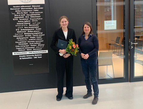 Dr. Sofia Meyer und Prof. Katharina von Kriegstein stehen nebeneinander vor einem Vorlesungsraum, Sofia hat einen Blumenstrauß in der den Händen