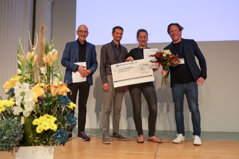 4 Personen stehen auf einer Bühne, 3 Männer und eine Frau.  Frau Christa Müller-Axt präsentiert den ihr überreichten GNP-Förder- und Nachwuchspreis.