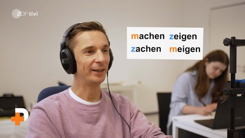 Ein Mann sitzt mit Kopfhörern vor einem PC. Neben ihm sind die Wörter machen, zeigen, zachen, meigen eingeblendet.