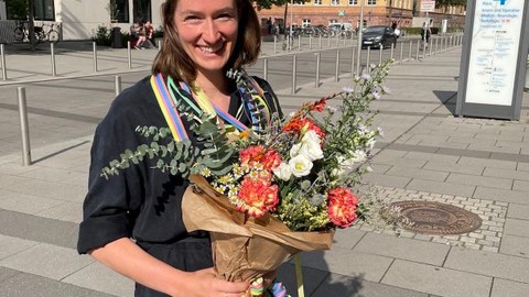 Leona Sureth steht im Freien vor der Universität Leipzig und trägt einen Doktorhut und hält einen Blumenstrauß in den Händen.