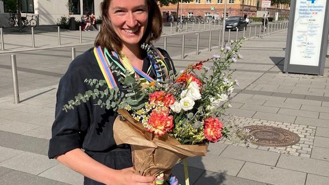 Leona Sureth steht im Freien vor der Universität Leipzig und trägt einen Doktorhut und hält einen Blumenstrauß in den Händen.