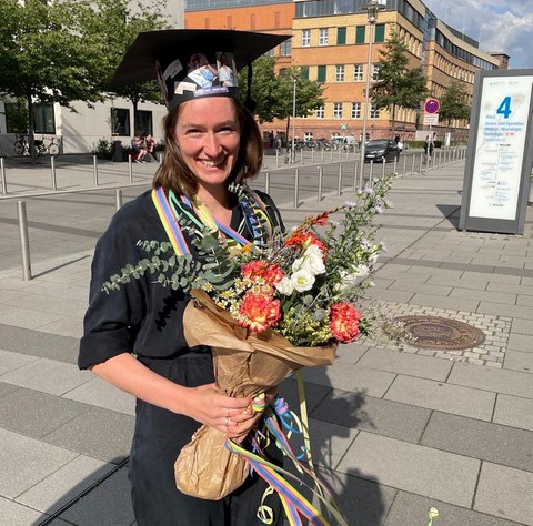 Leona Sureth steht im Freien vor der Universität Leipzig und trägt einen Doktorhut und hält einen Blumenstrauß in den Händen.