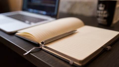 Foto: Arbeitsplatz mit Laptop, offenem Notizbuch mit Stift und einer Tasse im Hintergrund.