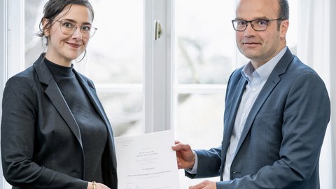 Frau Professorin Schiele bekommt von einem Mann ihre Ernennungsurkunde überreicht. Beide Personen sind förmlich gekleidet.