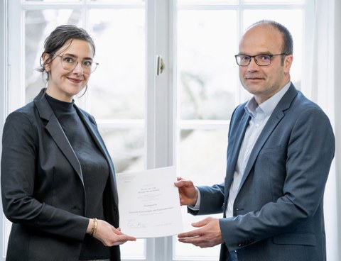 Frau Professorin Schiele bekommt von einem Mann ihre Ernennungsurkunde überreicht. Beide Personen sind förmlich gekleidet.