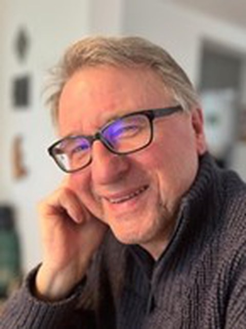 Portraitaufnahme von Claus Hertl, ein Mann mittleren Alters mit kurzem grauen Haar und einer dunklen, eckigen Brille, der freundlich in die Kamera lächelt.