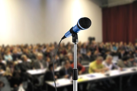 Das Foto zeigt ein Mikrofon, welches vor einem großen Publikum steht