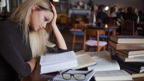 Auf dem Foto ist eine Person zu sehen. Sie sitzt an einem Tisch. Vor ihr liegen aufgeschlagene Bücher und Bücher auf einem Stapel. Ihr Kopf ist auf ihre Hände abgestützt.