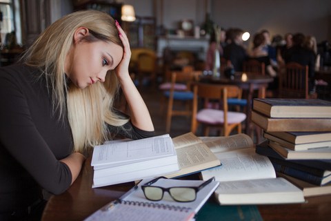 Auf dem Foto ist eine Person zu sehen. Sie sitzt an einem Tisch. Vor ihr liegen aufgeschlagene Bücher und Bücher auf einem Stapel. Ihr Kopf ist auf ihre Hände abgestützt.