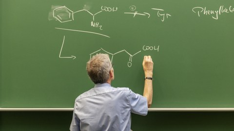 Das Foto zeigt einen Mann, der chemische Formeln auf eine Tafel schreibt.