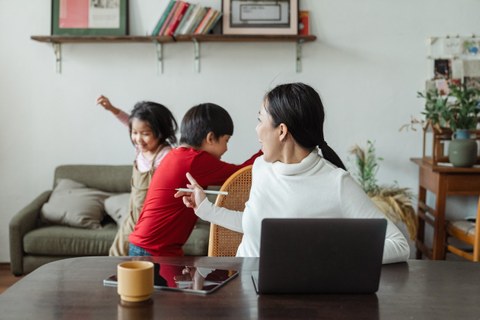 Frau am Laptop, im Hintergrund spielen zwei Kinder im Wohnzimmer