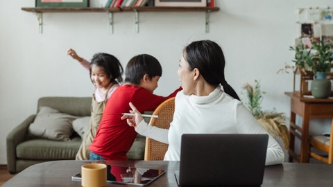 Frau am Laptop, im Hintergrund spielen zwei Kinder im Wohnzimmer