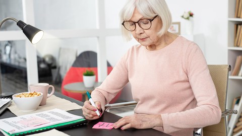 Eine ältere Dame mit Brille und weißem Haar sitzt an einem Bürotisch und und notiert etwas auf einem Klebezettel.