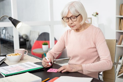 Eine ältere Dame mit Brille und weißem Haar sitzt an einem Bürotisch und und notiert etwas auf einem Klebezettel.