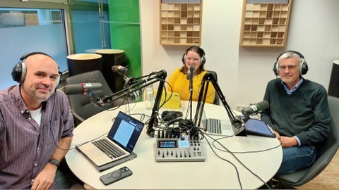 In einem Podcaststudio mit viel Equipment sitzen drei Menschen vor den Mikrofonen.
