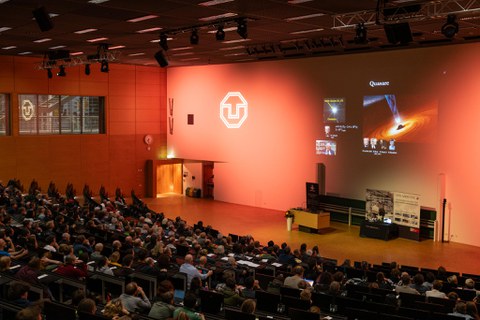 Foto von hinten über die Köpfe der Zuschauenden hinweg. Vorne sieht man die Powerpoint von Reinhard Genzel auf der er und eine Darstellung eines schwarzen Lochs ist