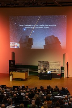 Foto von hinten über die Köpfe der Zuschauenden hinweg. Vorne sieht man die Powerpoint von Reinhard Genzel auf der er und ein Bild des "very large telescop"  zu sehen ist