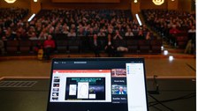 Foto vom Pult und des Laptops, über den Reinhard Genzel in den Audimax übertragen wird. Im Hintergrund sitzen sehr viele Interessierte Zuhörende.