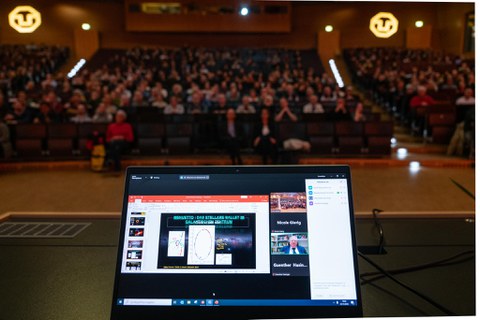 Foto vom Pult und des Laptops, über den Reinhard Genzel in den Audimax übertragen wird. Im Hintergrund sitzen sehr viele Interessierte Zuhörende.
