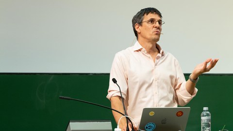 Physik Nobelpreisträger Didier Queloz am Pult vom Audimax der TU Dresden. Unter den vier Stickern auf seinem MacBook ist ein blauer mit einem Fisch. Um diesen steht: "JA zur Biodiversität" 