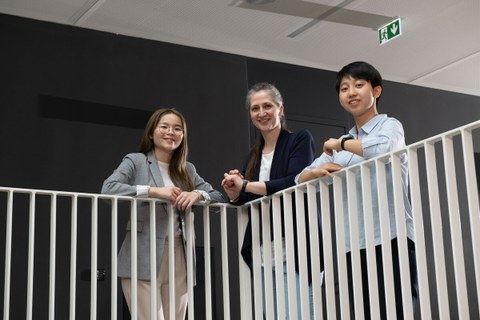 Foto von zwei taiwanesischen und einem deutschen Studierenden, die im Hermann-Krone-Bau an einer weißen Balustrade stehen und in die Kamera lächeln.
